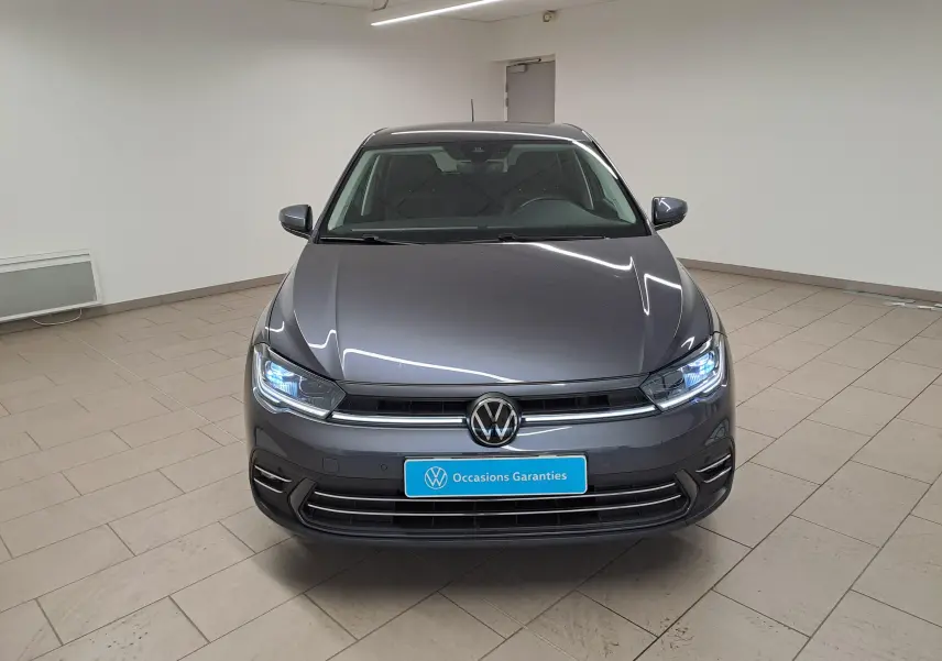Vue avant d'une Volkswagen Polo 2023 gris cendré avec phares allumés dans un showroom lumineux.