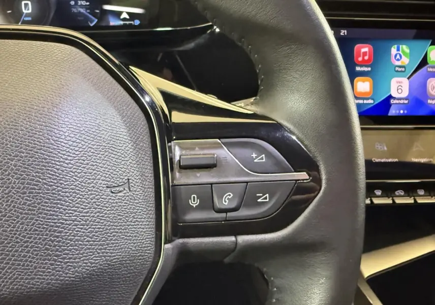 Gros plan sur le volant cuir noir de la Peugeot 308 2022 avec commandes vocales et écran tactile en arrière-plan.