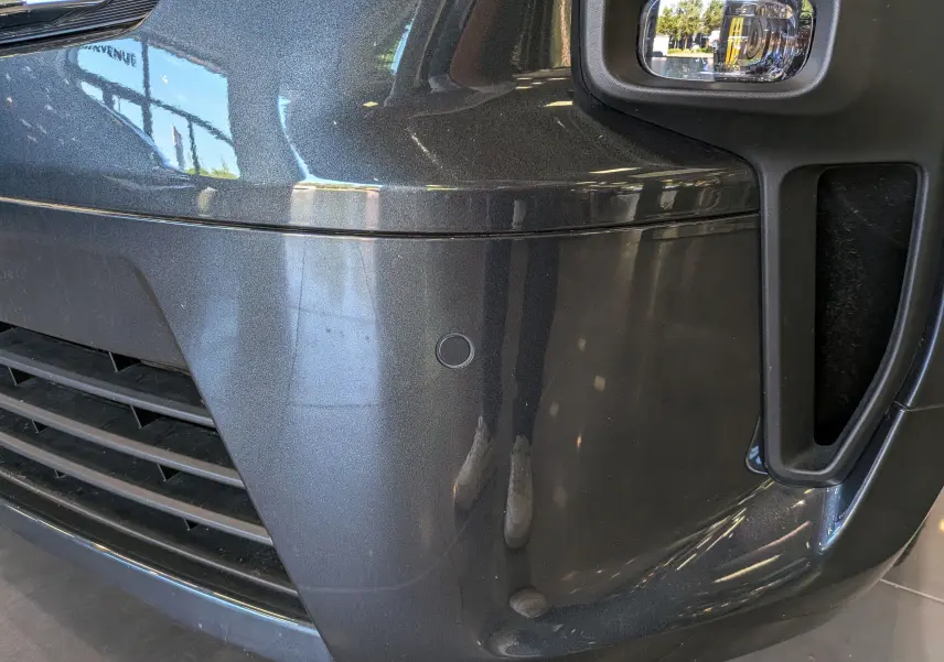 Gros plan sur le pare-chocs avant gris comète du Nissan Primastar Combi, avec feu antibrouillard et capteur de stationnement visible.