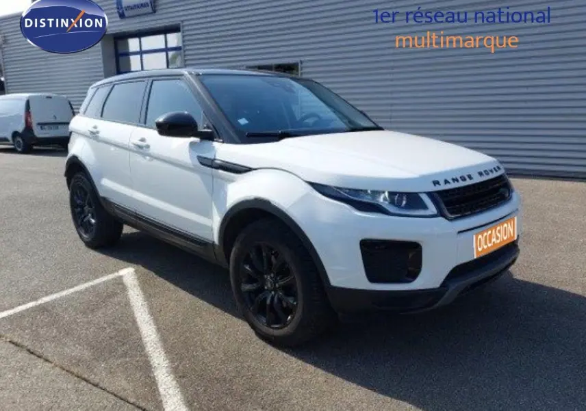 Range Rover Evoque blanc en 3/4 avant droit avec jantes noires, stationné devant un bâtiment industriel.