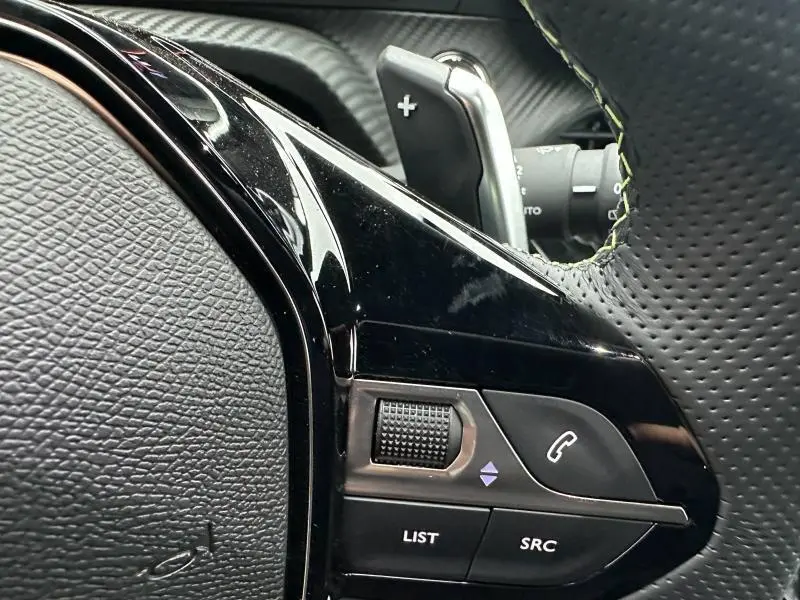 Gros plan sur les commandes noires du volant côté gauche du Peugeot 208 Hybrid 2025 avec surpiqûres vertes visibles.