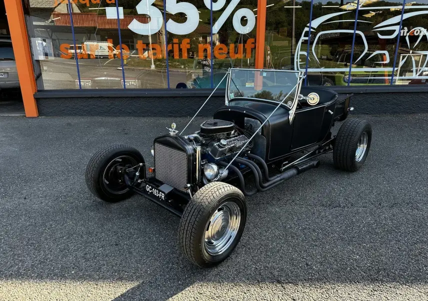 Ford T Hot Rod V8 noir vue 3/4 avant droit, capote manuelle baissée et moteur V8 apparent sur parking.