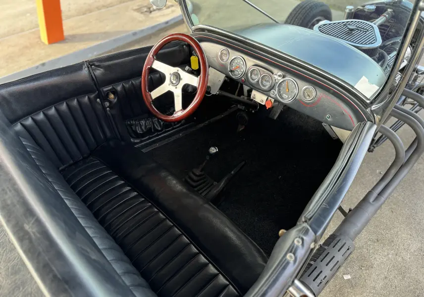 Intérieur noir cuir du Ford T Hot Rod 1923, vue plongeante sur banquette, volant bois et tableau de bord vintage.