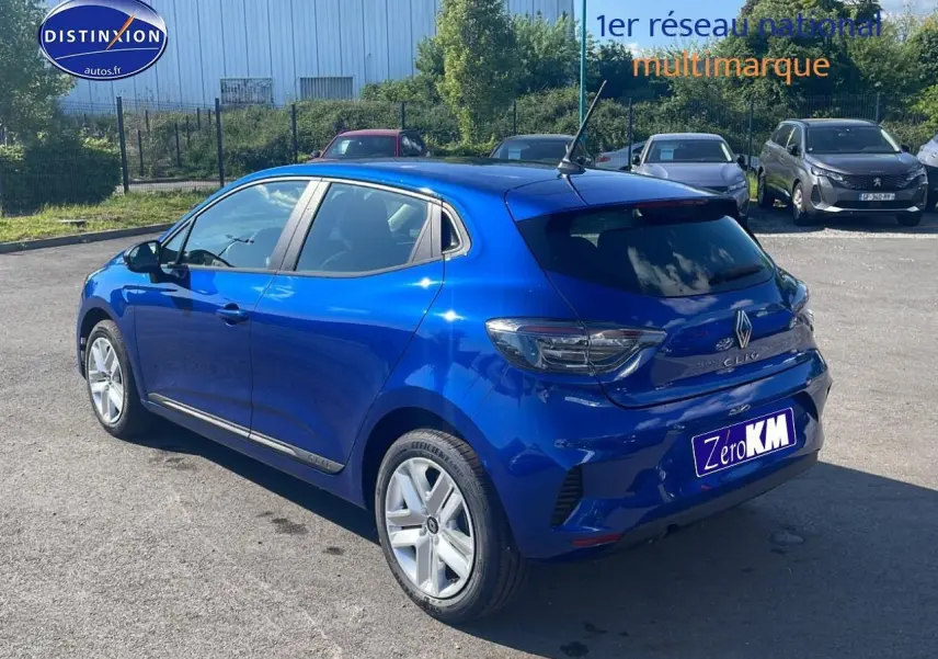 Renault Clio V bleu iron vue 3/4 arrière droit sur parking avec becquet et feux arrière LED teintés.