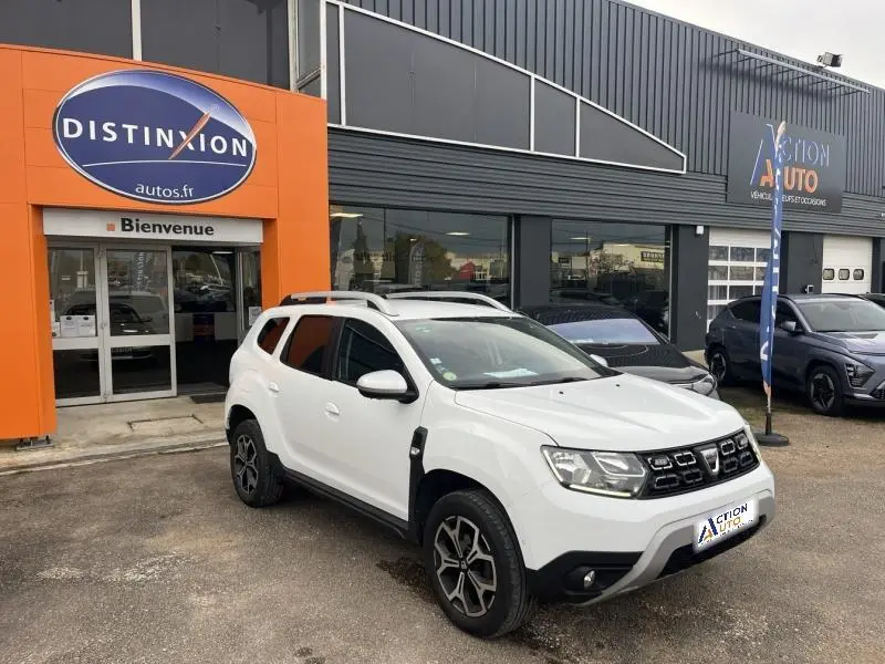 Dacia Duster blanc glacier en 3/4 avant droit avec jantes alliage et barres de toit chromées devant concession.