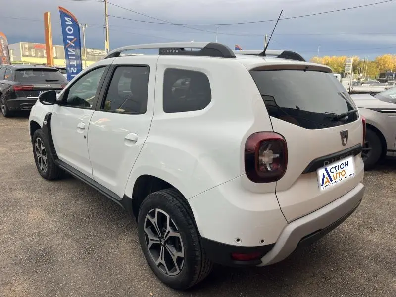 Vue 3/4 arrière droite du Dacia Duster blanc glacier avec jantes alliage et barres de toit chrome satiné.