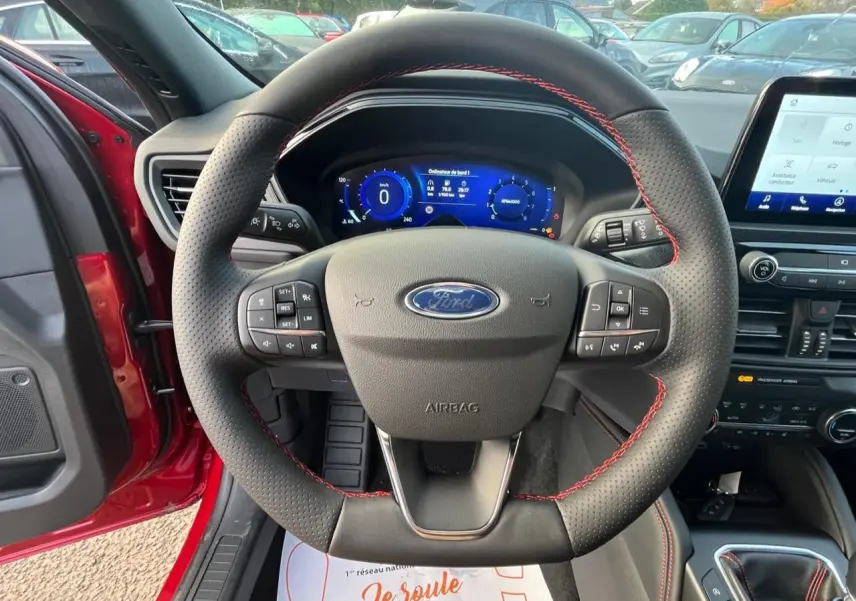 Vue intérieure centrée sur le volant perforé avec surpiqûres rouges du Ford Kuga rouge Lucifer, tableau de bord digital visible.