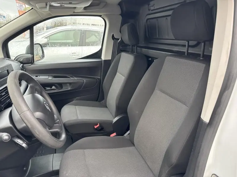 Vue intérieure côté conducteur du Citroën Berlingo Fourgon blanc, montrant les sièges tissu gris et le tableau de bord.