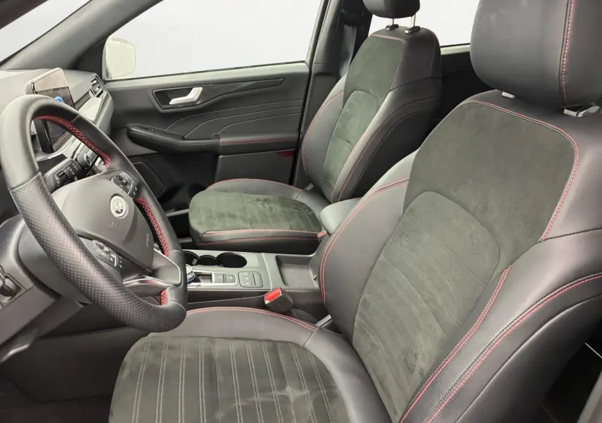 Vue intérieure côté conducteur du Ford Kuga 2024, sièges noirs en cuir et alcantara avec surpiqûres rouges, volant multifonction.