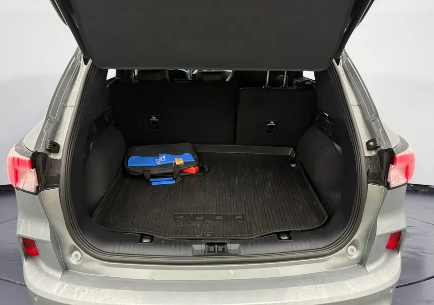Coffre ouvert vu de l'arrière d'un Ford Kuga gris 2024, avec tapis noir et trousse de secours bleue visible.