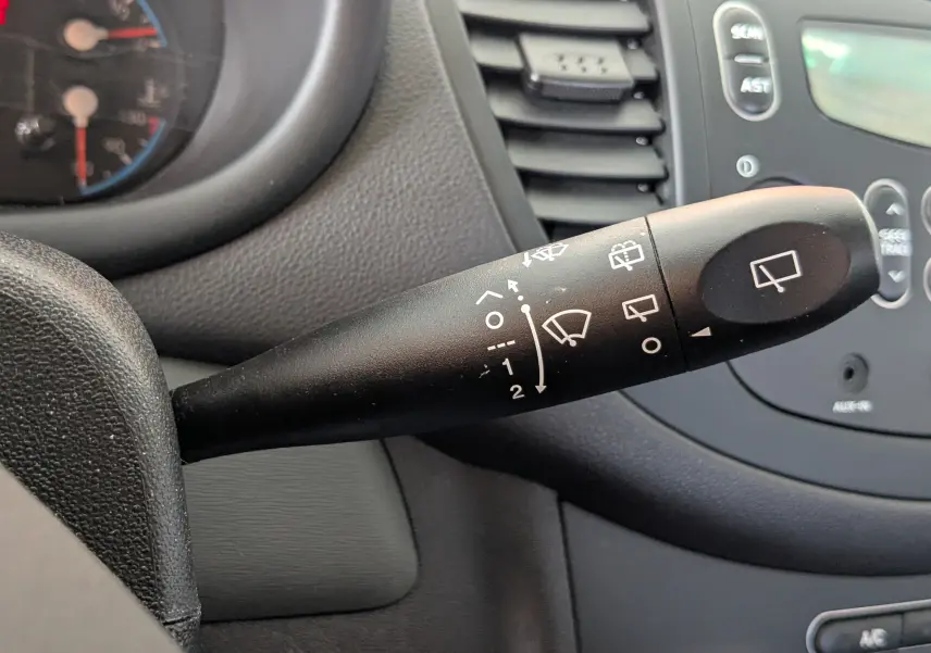 Gros plan sur la commande d'essuie-glace noire à l'intérieur d'une Hyundai i10 gris clair de 2010, tableau de bord en arrière-plan.