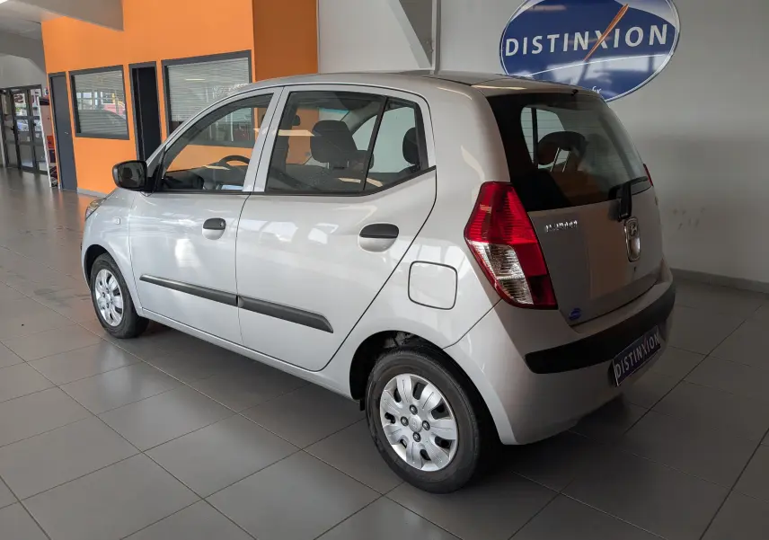 Vue 3/4 arrière droite d'une Hyundai i10 gris clair 5 portes, avec poignées noires et jantes acier en intérieur showroom.