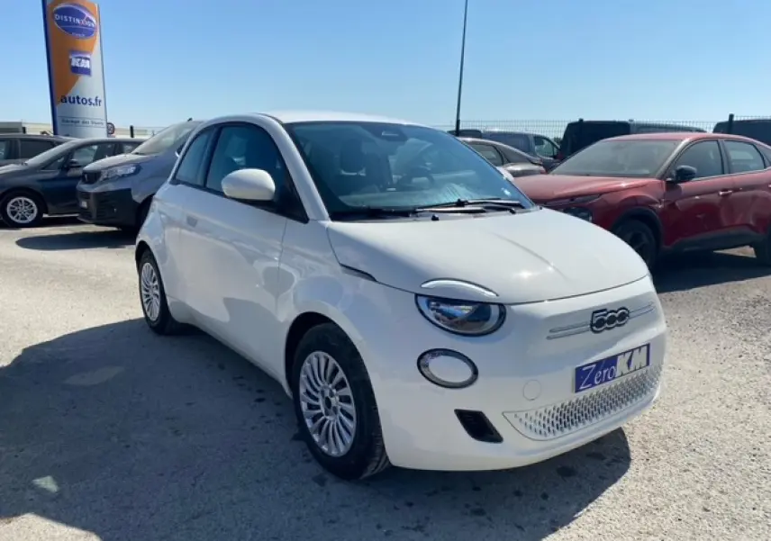 Vue 3/4 avant droite d'une Fiat 500 électrique blanche métallisée 2022 avec logo 500 et plaque Zero KM visible.