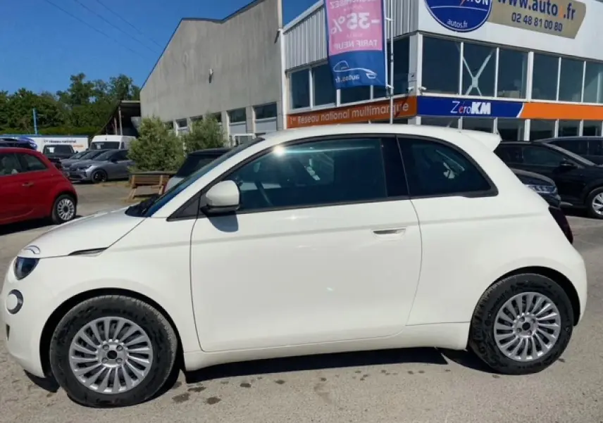 Vue de profil côté gauche d'une FIAT 500 électrique blanche métallisée, stationnée devant un bâtiment commercial.