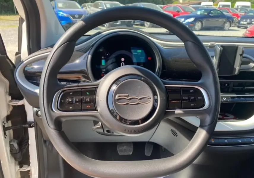 Vue intérieure centrée sur le volant noir de la Fiat 500 électrique blanche, avec tableau de bord numérique et support smartphone visible.