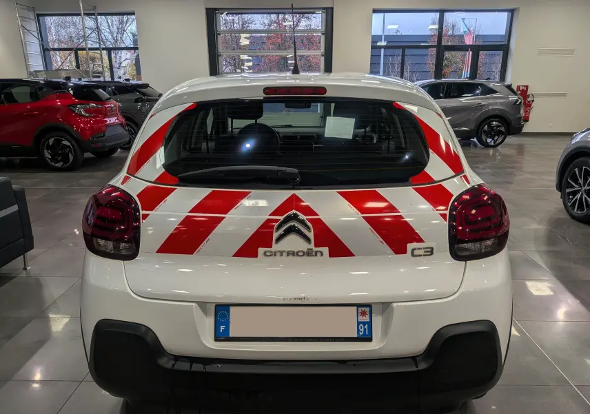 Vue arrière d'une Citroën C3 Société blanc banquise avec bandes rouges réfléchissantes en diagonale dans un showroom.
