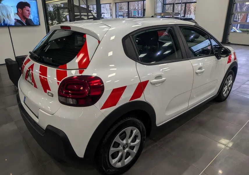 Vue 3/4 arrière droite d'une Citroën C3 Société blanche avec bandes rouges diagonales et feux arrière foncés.