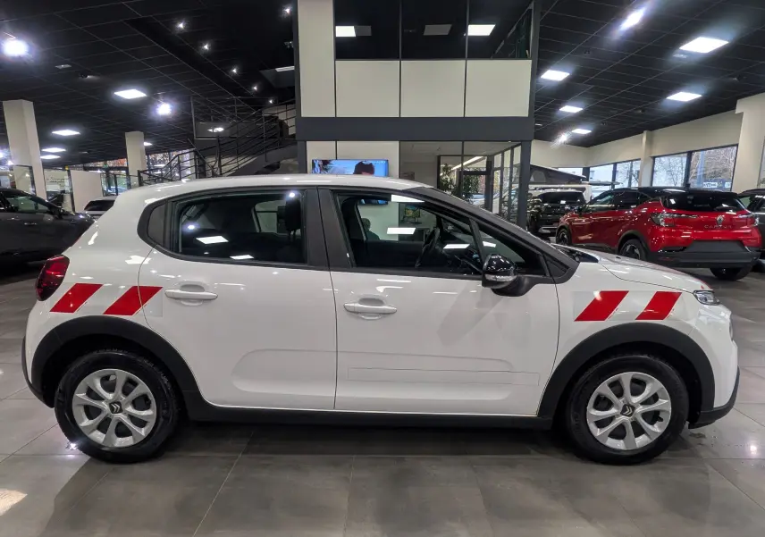 Profil côté gauche d'une Citroën C3 Société blanche avec bandes rouges diagonales sur les ailes avant et arrière, en showroom.