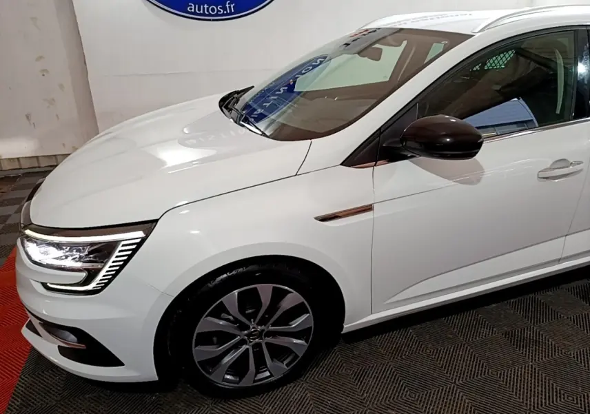Vue 3/4 avant droit d'une Renault Mégane Techno TCe 140 blanche avec rétroviseur noir et feux LED allumés.