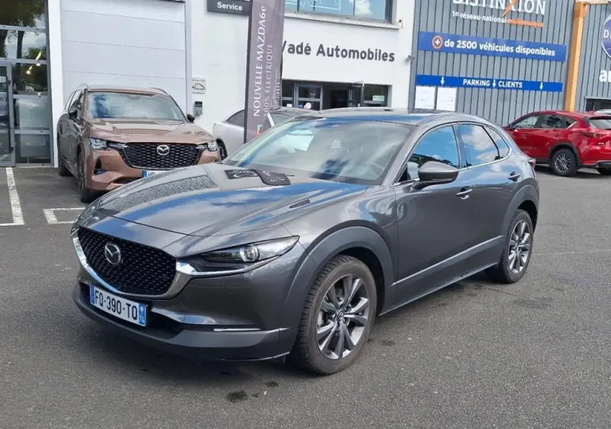 Mazda CX-30 gris en 3/4 avant droit sur parking devant concession avec jantes aluminium visibles.