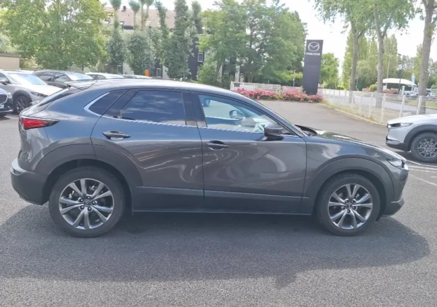 Vue de profil côté gauche d'un Mazda CX-30 gris avec vitres surteintées et jantes aluminium, stationné en extérieur.
