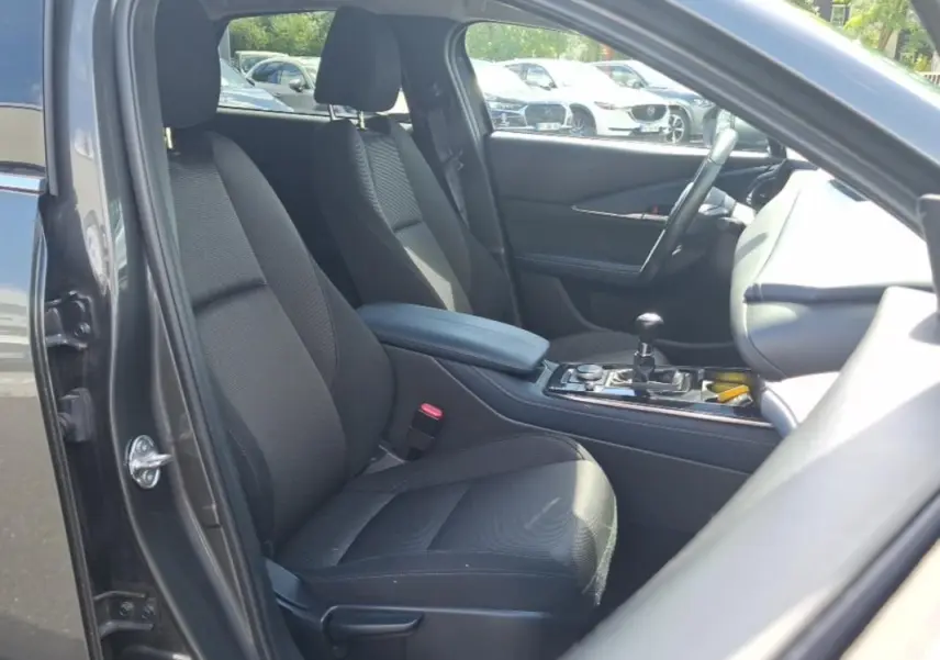 Vue intérieure côté conducteur du Mazda CX-30 gris, montrant les sièges noirs, accoudoir central et volant multifonctions.