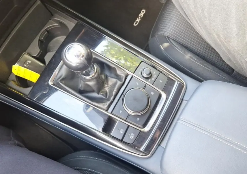 Vue plongeante sur la console centrale noire brillante du Mazda CX-30 avec levier de vitesse manuel et accoudoir gris.