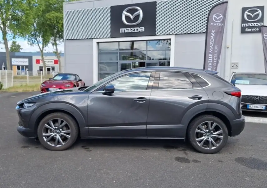 Vue de profil côté gauche d'un Mazda CX-30 gris stationné devant un concessionnaire Mazda avec jantes aluminium.