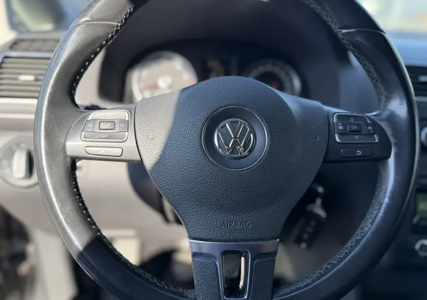 Gros plan sur le volant en cuir noir du Volkswagen Touran gris 2012, avec commandes intégrées visibles.