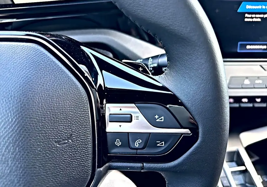 Gros plan sur les commandes du volant multifonction noir de la Peugeot 308 gris selenium, avec écran tactile en arrière-plan.