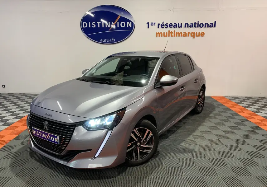 PEUGEOT 208 gris vue 3/4 avant droit, phares LED allumés, sur sol à damiers gris et orange en intérieur showroom.