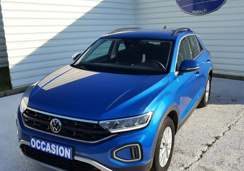 Volkswagen T-Roc bleu en 3/4 avant droit, avec calandre noire et feux LED, stationné devant un mur blanc.