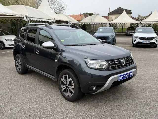 Vue 3/4 avant droit d'un Dacia Duster gris comète 2022 avec barres de toit et jantes alliage distinctives.