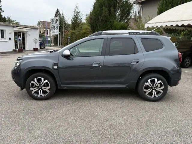 Vue de profil côté gauche d'un Dacia Duster 2022 gris comète avec jantes alliage et barres de toit noires.