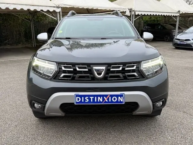 Vue avant d'un Dacia Duster 2022 gris comète avec calandre noire et feux LED allumés sur un parking.