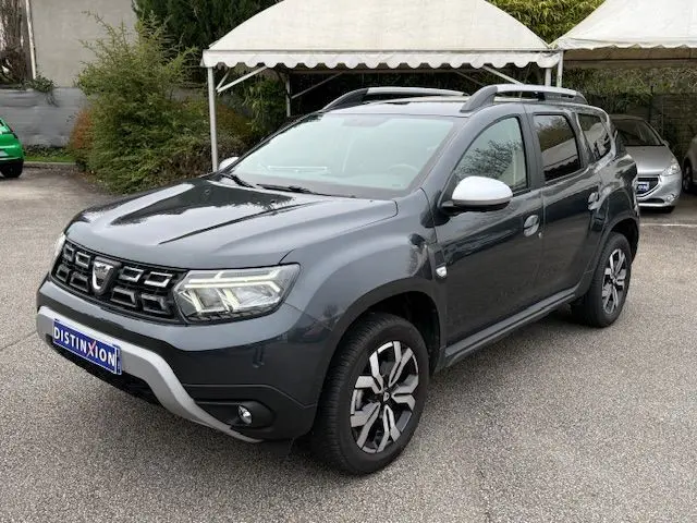 Dacia Duster gris comète en 3/4 avant droit, avec barres de toit argentées et jantes alliage distinctives.