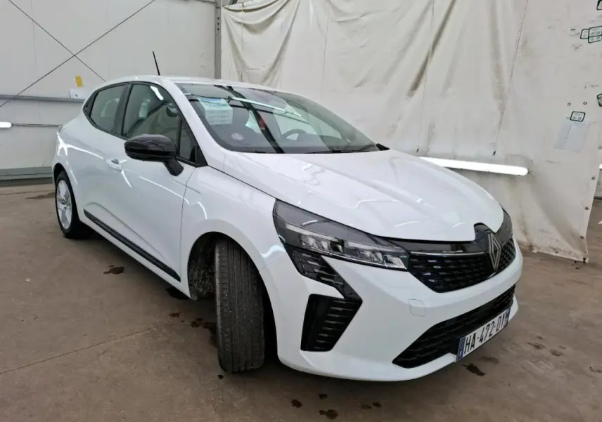 Renault Clio blanche vue 3/4 avant droit, avec calandre noire et feux LED distinctifs, stationnée en intérieur.
