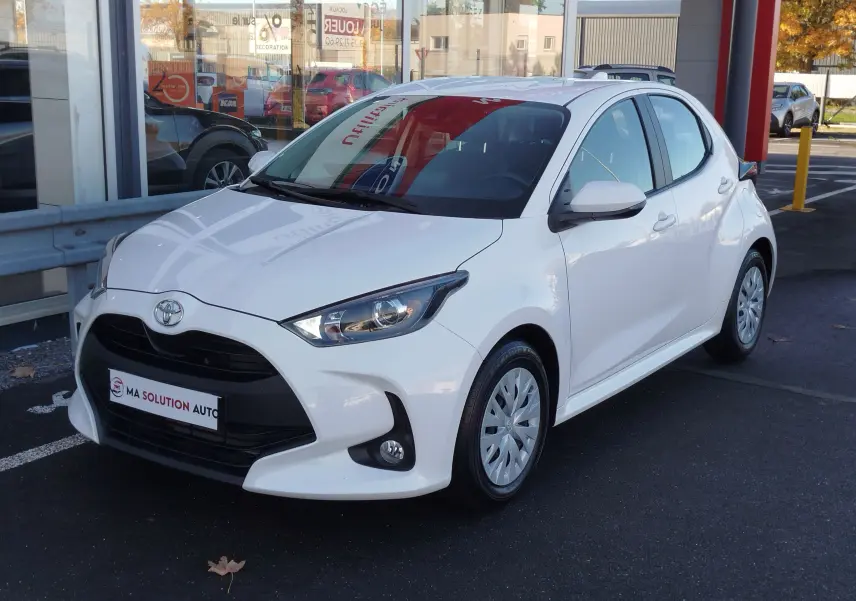Toyota Yaris blanc pur vue 3/4 avant droit, avec calandre noire et feux avant LED visibles, stationnée devant un showroom.