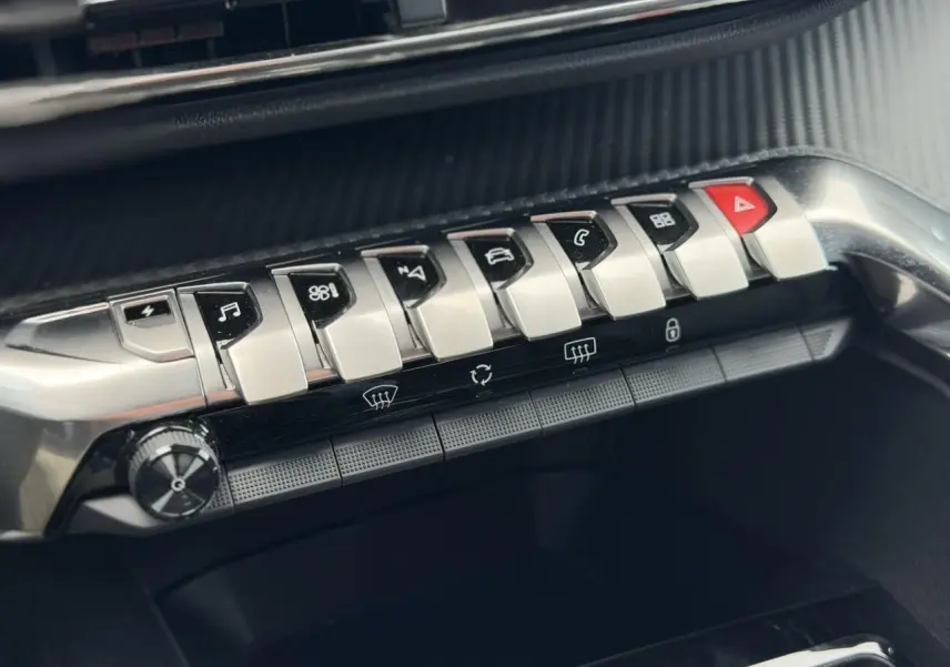 Gros plan sur la rangée de boutons métalliques du tableau de bord du Peugeot 3008 Plug-in Hybrid noir, avec touches fonctionnelles et alerte.