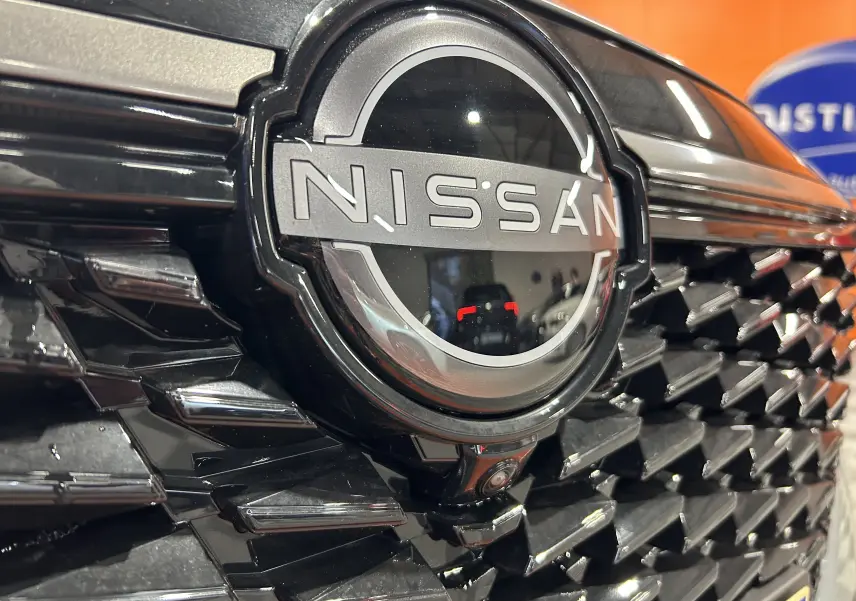 Gros plan sur la calandre noire brillante avec logo Nissan et caméra avant du Qashqai gris céramique 2025.