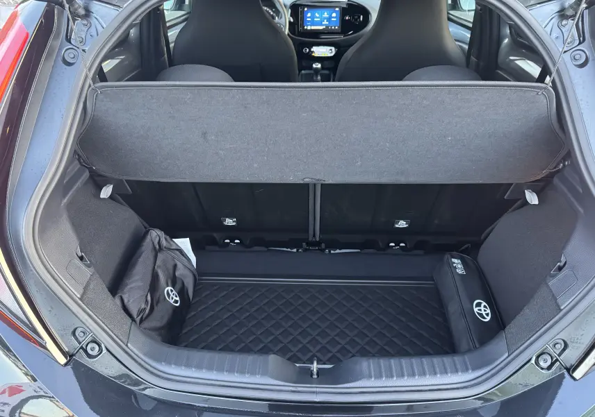 Coffre ouvert vu de l’arrière d’une Toyota Aygo X bleue, montrant l’espace de rangement et l’intérieur avec écran tactile.