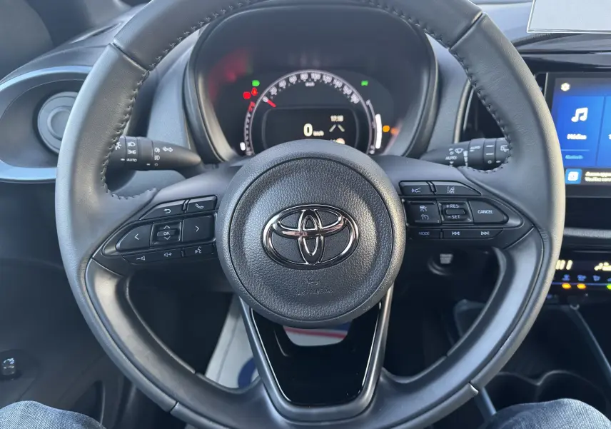 Vue intérieure centrée sur le volant noir de la Toyota Aygo X 2025, avec tableau de bord et commandes au volant visibles.