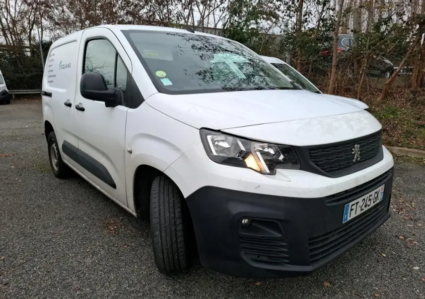 Peugeot Partner blanc vue 3/4 avant gauche avec phare allumé et pare-chocs noir sur parking extérieur.