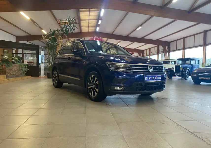 Volkswagen Tiguan bleu vert 2020 vu en 3/4 avant droit dans un showroom avec voitures anciennes en arrière-plan.