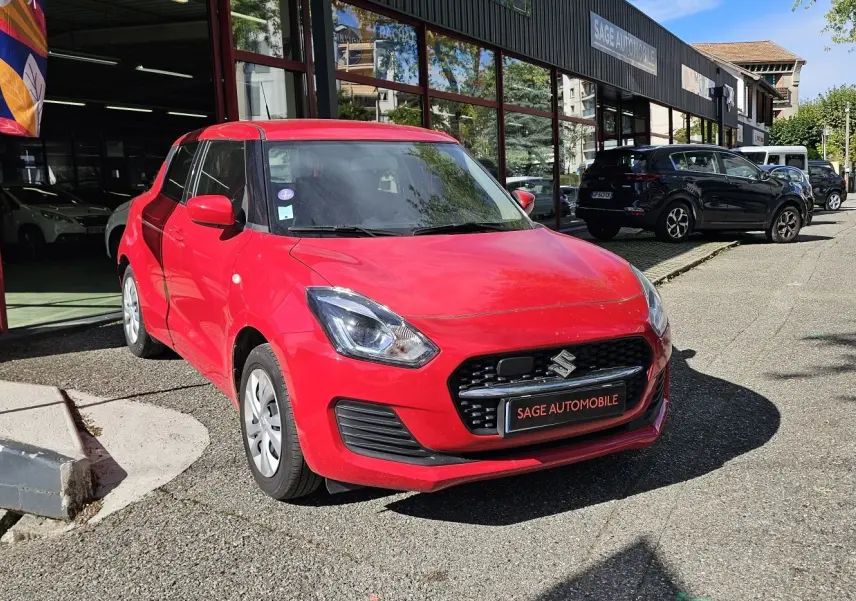 Suzuki Swift 2023 rouge fervent vue 3/4 avant garée devant un garage avec plaque Sage Automobile.
