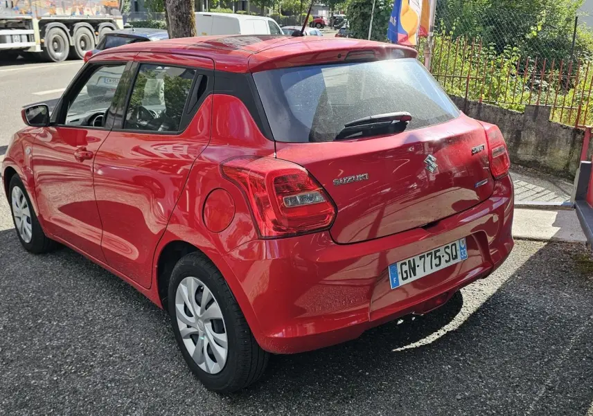 Vue 3/4 arrière droite d'une Suzuki Swift 1.2 Dualjet Hybrid 2023 en rouge Fervent, avec hayon et feux arrière visibles.