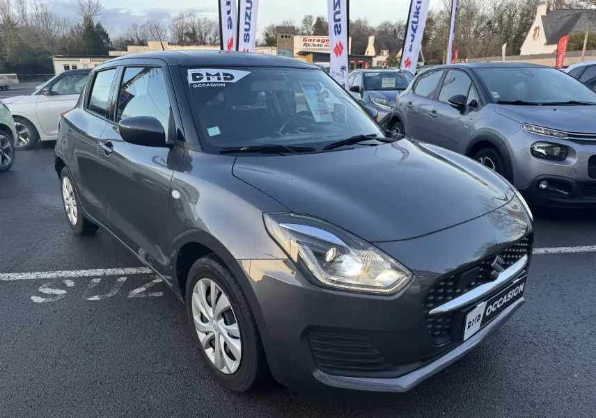 Suzuki Swift 1.2 Dualjet Hybrid gris vue 3/4 avant gauche, avec phares allumés et autocollant "Occasion" sur le pare-brise.