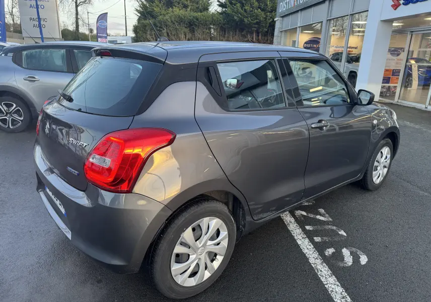 Vue 3/4 arrière droite d'une Suzuki Swift 1.2 Dualjet Hybrid gris métallisé, garée devant un concessionnaire.