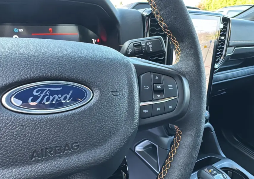Gros plan sur le volant cuir noir avec surpiqûres orange du Ford Ranger PHEV 281 Wildtrak, tableau de bord et écran tactile visibles.