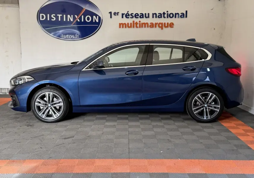 BMW Série 1 116i Business Design bleu vue de profil côté gauche dans un showroom avec sol gris et orange.